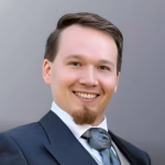 Foto_RobertSkawinski Porträt von Robert Skawinski, ein lächelnder Mann im Anzug mit Krawatte vor neutralem Hintergrund.