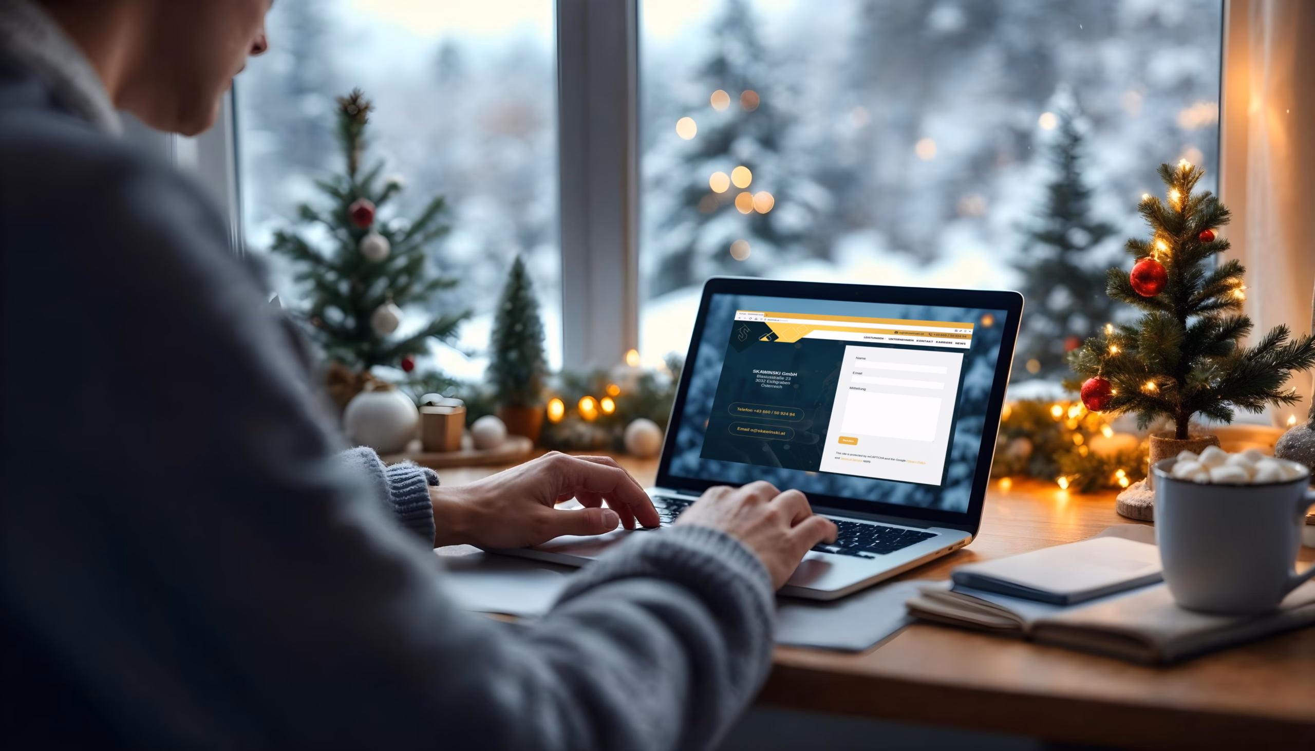Weihnachten_kontakt Eine Person tippt an einem festlich dekorierten Schreibtisch auf einem Laptop. Im Hintergrund sind ein Fenster mit Schneelandschaft und weihnachtlicher Dekoration zu sehen.