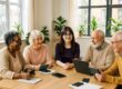 Katharina Grünwald (Mitte) leitet einen Digital-Workshop für vier Seniorinnen in einem hellen Raum. Die Teilnehmerinnen sitzen gemeinsam an einem Holztisch und arbeiten mit Tablets und Smartphones. Die freundliche Atmosphäre zeigt die praxisnahe Vermittlung digitaler Kompetenzen im Rahmen des Projekts Digital Überall Plus.
