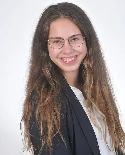 Porträt von Daniela Söldner, eine lächelnde junge Frau mit langen welligen Haaren und feiner Brille, im dunkelblauen Blazer über weißem Oberteil vor hellem, neutralem Hintergrund.