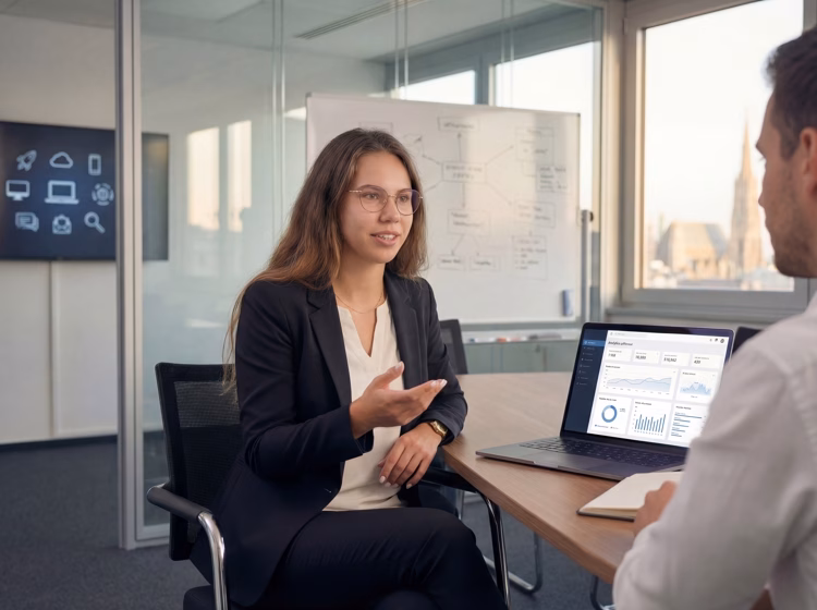 IT consultant Daniela Söldner advising a client in a modern Vienna office, with analytics dashboard on laptop and IT infrastructure diagram on whiteboard, Vienna skyline visible through window